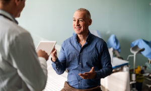 A smiling man in a blue shirt engages with a healthcare professional holding a tablet in a clinical setting.