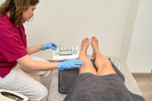 A technician in scrubs uses a device on a patient's legs, which are covered with a dark towel. The room is well-lit.