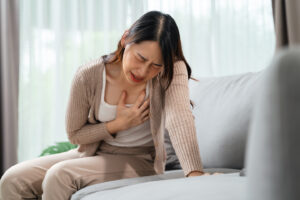 A woman sits on a couch, clutching her chest with a pained expression, suggesting distress or discomfort.
