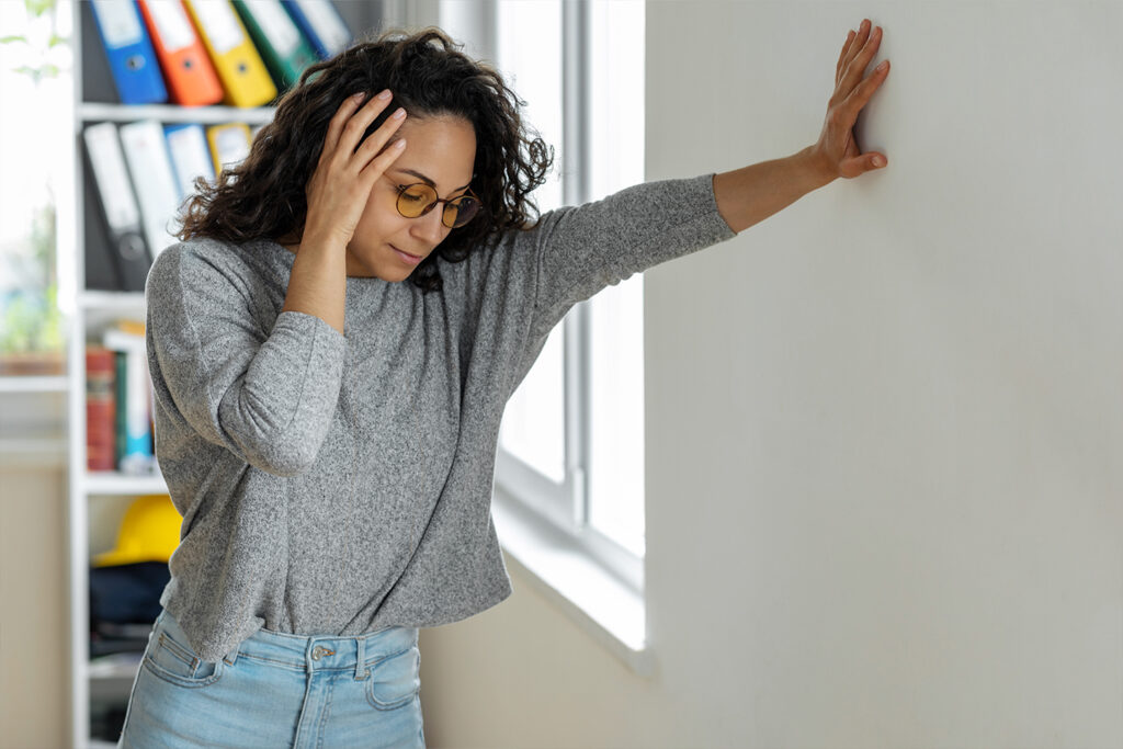 A woman with curly hair and glasses leans against a wall, looking stressed while holding her head in her hand.